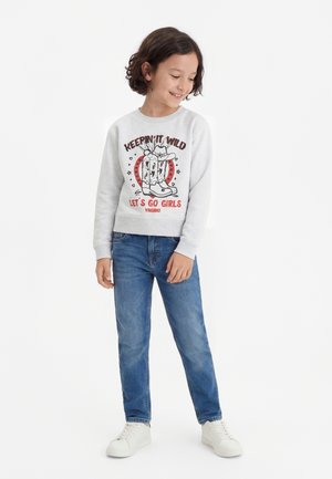 Smilende barn med kort krøllete hår iført en lys genser med cowboyboots-trykk og jeans, stående foran en hvit bakgrunn.