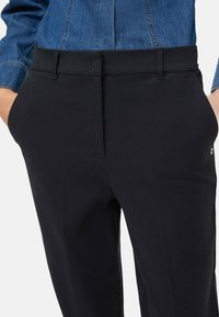 Pantaloni neri con una texture liscia, dotati di tasche laterali e una cintura sagomata. Un delicato dettaglio metallico è visibile sulla tasca.