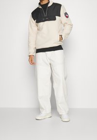 Krämpullover i fleece med svart panel, halvdragkedja och logotyp; i kombination med löst sittande vita byxor och vita sneakers.