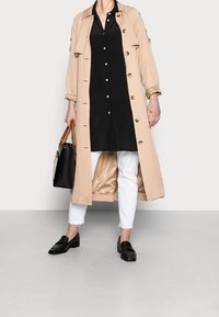 Cappotto lungo beige con bottoni grandi, indossato sopra un vestito camicia nero. Jeans bianchi alla caviglia e mocassini in pelle nera completano il look.
