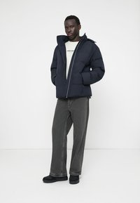 Marineblauer Pufferjacke mit Reißverschluss und Kapuze, ausgestattet mit einer glatten Textur. Kombiniert mit grauen Jeans und schwarzen Schuhen.