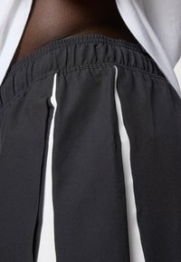 Detailní záběr elastického pásku na černých kalhotách s bílými vertikálními pruhy, které jsou nošeny s bílou košilí, s důrazem na texturu látky a střih.