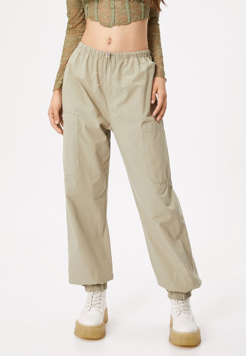 Pantalones cargo de color beige claro con cintura elástica, dos bolsillos laterales y puños elásticos. Combinados con un top corto de encaje semitransparente y botas de platform blancas.