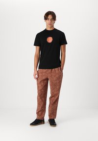 Black short-sleeve t-shirt with a circular multicolored graphic; brown patterned pants with intricate floral designs; black sneakers.