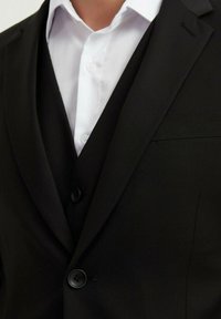 Veste de costume noire en tissu texturé, avec revers crantés, fermeture à un bouton et une chemise blanche en dessous.