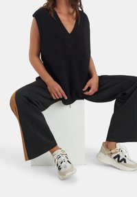 Haut sans manches en maille bleu marine avec un col en V, associé à un pantalon fendu noir et marron, et des baskets beiges avec un lacet et un design en maille.