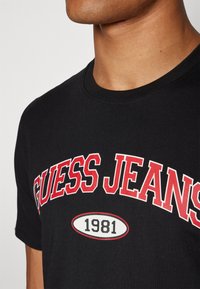 Juodas medvilninis marškinėliai su „GUESS JEANS“ išryškintomis raudonomis raidėmis ir baltu ovuliu, kuriame spausdinamas „1981“ žemiau teksto. Klasikinis apvalus kaklo dizainas.