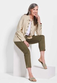 Veste beige à boutons sur une chemise blanche, pantalon olive, et sandales à glissière vert clair. Caractéristiques : coupe décontractée et couture visible.