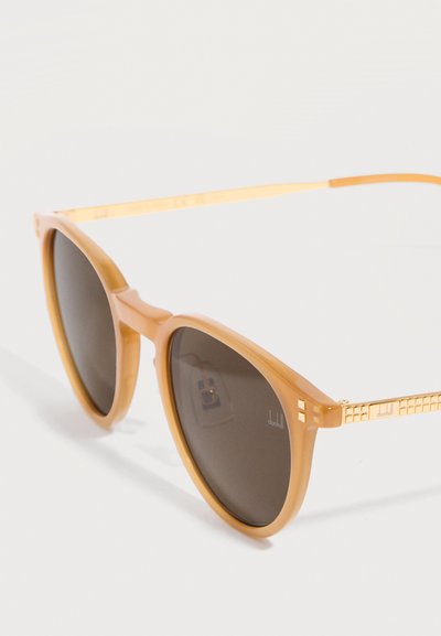 Γυαλιά ηλίου με στρογγυλό σκελετό, διαφανείς ανοιχτό καφέ περιμετρικές άκρες και χρυσαφένιους μεταλλικούς βραχίονες με υφή, με σκουρόχρωμους φακούς.