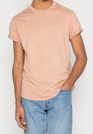Mann trägt ein hellrosa T-Shirt mit aufgekrempelten Ärmeln und hellblauen Jeans, eine Hand in der Tasche, vor einfachem Hintergrund.
