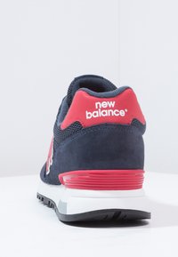 Vue arrière d'une chaussure de sport New Balance bleu marine et rouge avec une semelle blanche sur une surface blanche.