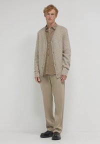 Cardigan beige lavorato a maglia con tessuto testurizzato, bottoni in legno e polsini a coste, indossato su una camicia marrone con bottoni e pantaloni beige. Scarpe nere.