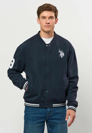 Młody mężczyzna nosi granatową kurtkę typu varsity z białymi paskami na mankietach i dole, białe logo gracza polo oraz niebieskie dżinsy.