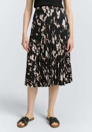 Woman wearing a black and white patterned pleated midi skirt, black top, and black open-toe slide sandals, standing on a plain background.