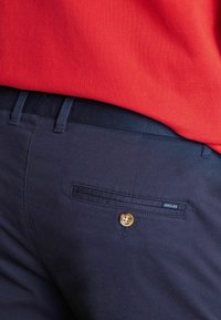 Marineblaue Hose mit einer hinteren Tasche mit Knopf und kleinem "Joules"-Etikett, getragen mit einem in die Hose gesteckten roten, strukturierten Hemd.