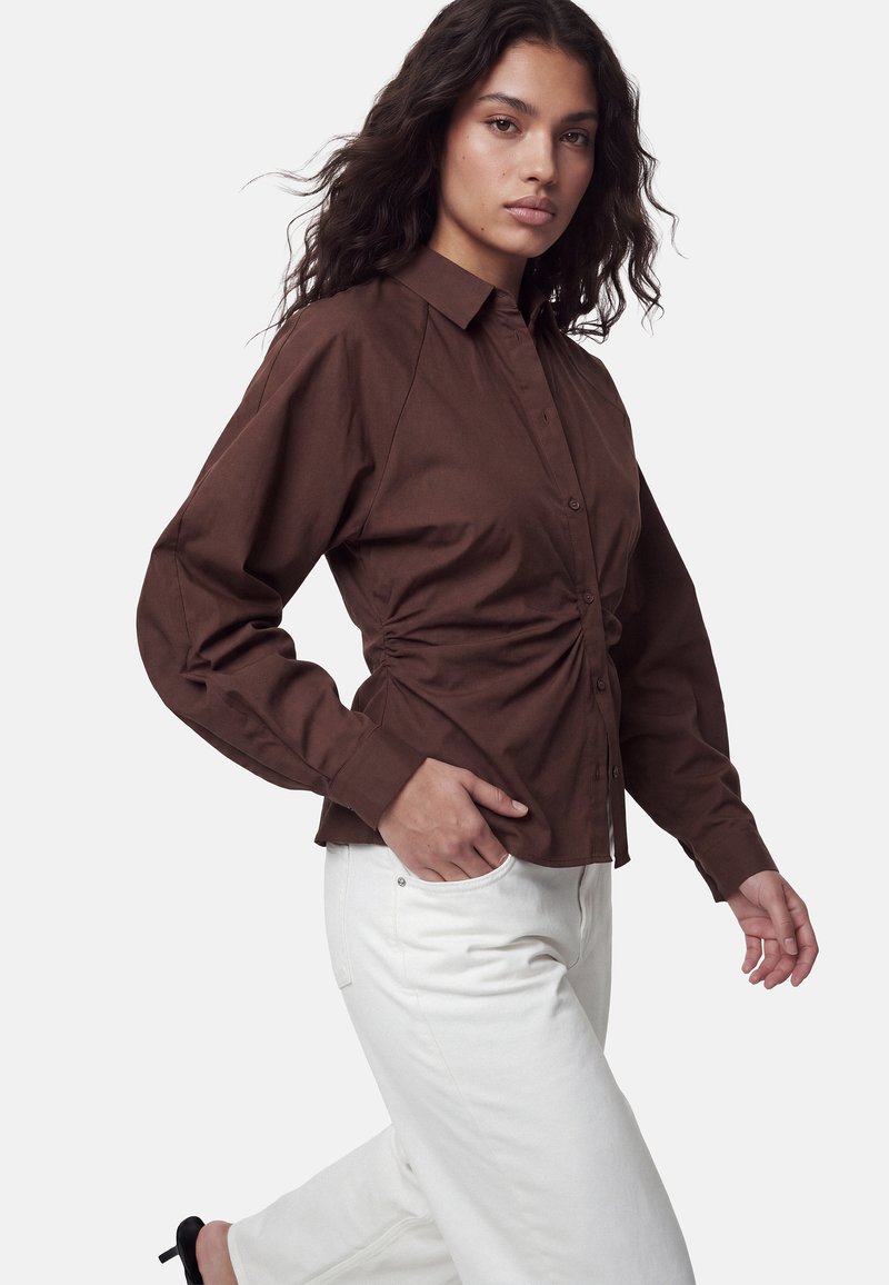 Femme portant une chemise marron foncé boutonnée et un pantalon blanc, posant avec une main dans la poche et légèrement de profil.