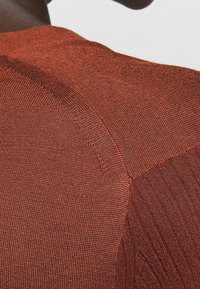 Sweter w kolorze rdzy z dzianiny, o fakturowanej tkaninie, z prążkowanymi detalami na rękawach i gładkimi sekcjami. Zbliżenie na plecy.
