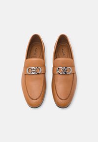 Mocassins en cuir tan avec un bout arrondi, dotés d'une boucle double-cercle en argent sur une sangle tan et d'un surfilage contrastant le long des bords.