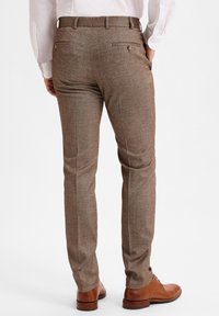 Braune, strukturierte Hose mit tailored Fit, ausgestattet mit Gesäßtaschen und versteckten Knöpfen. Kombiniert mit braunen Lederschuhen.