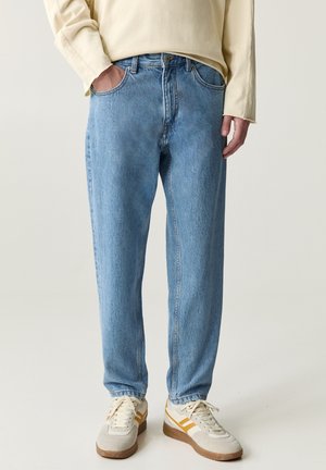 Person wearing light blue straight-fit jeans, cream long-sleeve shirt, and beige sneakers with yellow stripes on a plain background.