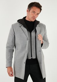 Šedý kabát z vlněné směsi s černou kapucí na zip, dvěma předními kapsami a zapínáním na knoflíky. Hladká textura s vykrojeným střihem.