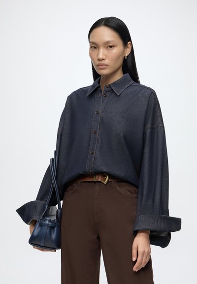 Femme aux cheveux noirs lisses portant une chemise en jean sombre oversize, un pantalon marron, et tenant un petit sac à main bleu foncé sur fond blanc uni.