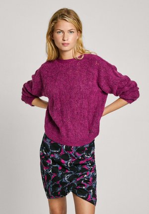 Fuchsia gebreide trui met ribbelkraag en oversized mouwen, gecombineerd met een zwarte rok met een meerkleurig bloemenpatroon.
