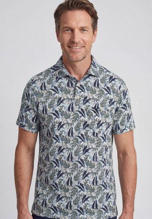 Man glimlacht, draagt een kortärmelig overhemd met kraag met een blauw-groen bladmotief op een lichte achtergrond, staand voor een effen achtergrond.