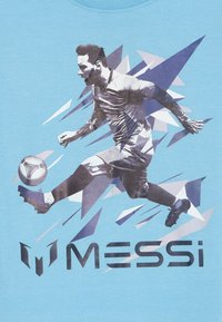 Šviesiai mėlynas marškinėlis su grafika, vaizduojančia futbolininką judesyje, spirdamas kamuolį, su kampuotais raštais violetinės ir pilkos spalvų. Po juo spausdinta „MESSI“. 