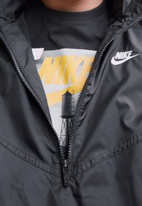 Henkilö pukeutuu mustaan Nike-takkiin, joka on puoliksi auki, paljastaen mustan Nike-t-paidan, jossa on keltainen logo ja vesitorni-grafiikka.