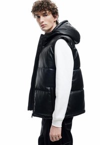 Schwarze gepolsterte Weste mit Kapuze, aus glänzendem Material. Sie verfügt über Seitentaschen, einen Reißverschluss und ein weißes Langarmshirt darunter.