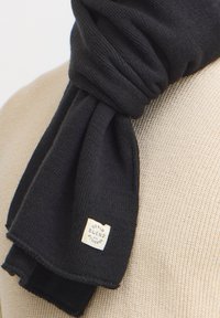 Sciarpa nera lavorata a maglia con una finitura testurizzata. Presenta un'etichetta con la scritta "Denim Blend with Attitude". La sciarpa è delicatamente appoggiata su uno sfondo neutro.