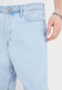 Lys blå denimjeans med rett snitt, synlig søm og framlommer; metallknapp og niterdetaljer. Glatt tekstur.