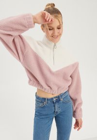 Femme debout, yeux fermés et souriante, portant un jean bleu taille haute et un pull zippé duveteux rose et blanc.