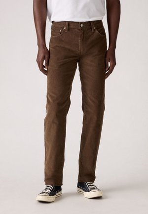 Mężczyzna w brązowych spodniach sztruksowych, białej koszulce i czarnych tenisówkach stoi na jednolitym tle.