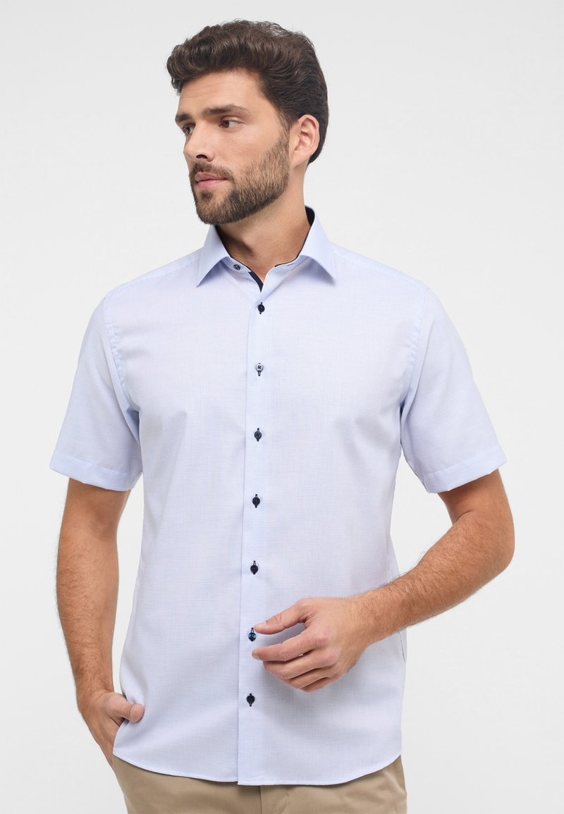 Camicia azzurra a maniche corte con bottoni, con un motivo testurizzato, bottoni blu scuro e colletto a punta. Il tessuto appare liscio e leggero.