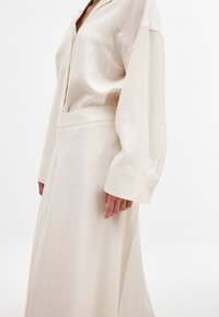 Beige långärmad klänning med krage, med ett bälte i midjan och mjukt, texturerat tyg. Ärmarna har knappmanchetter och en avslappnad passform.