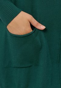 Tessuto lavorato a maglia verde scuro con un polsino a coste e una tasca frontale. La tasca ha un'apertura arrotondata, evidenziando la morbidezza della texture.