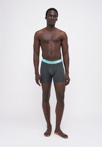 Sive bokserice Nike z turkizno elastiko, ki nudijo tesno prileganje in gladko teksturo. Model stoji bos, roke sproščene ob straneh.