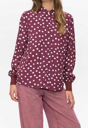 Kvinde iført en langærmet bordeaux bluse med hvide ujævne polkaprikker og lyserøde bukser med vide ben, stående med den ene hånd på hoften.