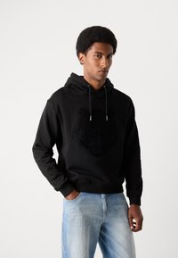 Hoodie nera con un emblema di leone in rilievo e testurizzato sul davanti. Presenta un cappuccio con cordini e polsini a coste. Indossata con jeans azzurri chiari.