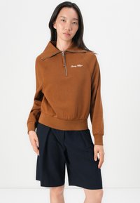 Brown zip-up sweatshirt with a large collar, ribbed hem, and embroidered logo. Paired with dark navy shorts. Overall relaxed fit.