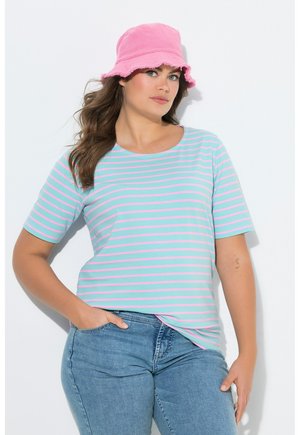 Vrouw met een roze bucket hat, turquoise en roze gestreept T-shirt en een blauwe jeans, poserend met één hand in de zak tegen een witte achtergrond.
