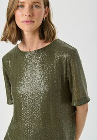 Top de culoare verde măsliniu, cu paiete, cu mâneci scurte și decolteu rotund, având un model texturat și un finisaj strălucitor care reflectă lumina.