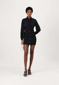 Minidress nero con maniche lunghe, chiusura con bottoni frontale, due tasche anteriori e vita aderente, abbinato a sandali neri e a una piccola borsa bianca.