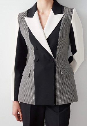 Schwarz-weißer zweireihiger Blazer mit strukturierten Musterpaneelen und kontrastierenden Revers, kombiniert mit schwarzen Hosen.