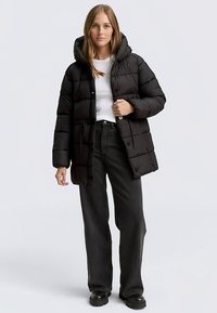 Schwarze, gesteppte Steppjacke mit Kapuze, mit Knöpfen vorne und seitlichen Taschen. Wird über einem weißen Hemd und einer schwarzen Hose getragen.