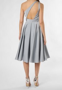 Žena v stříbrné saténové midi šatech s jedním ramínkem a odhaleným zády, doplněná stříbrnými vysokými podpatky, stojící na bílém pozadí.