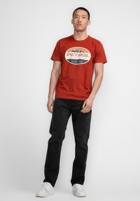 Camiseta de algodón roja con un gráfico circular en blanco y negro que dice "PETROL." Combinada con jeans negros y zapatillas deportivas blancas.