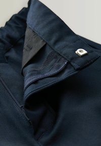Tissu bleu foncé avec une texture lisse. Présente une fermeture éclair et des éléments en métal. La taille a une finition soignée et des coutures détaillées.